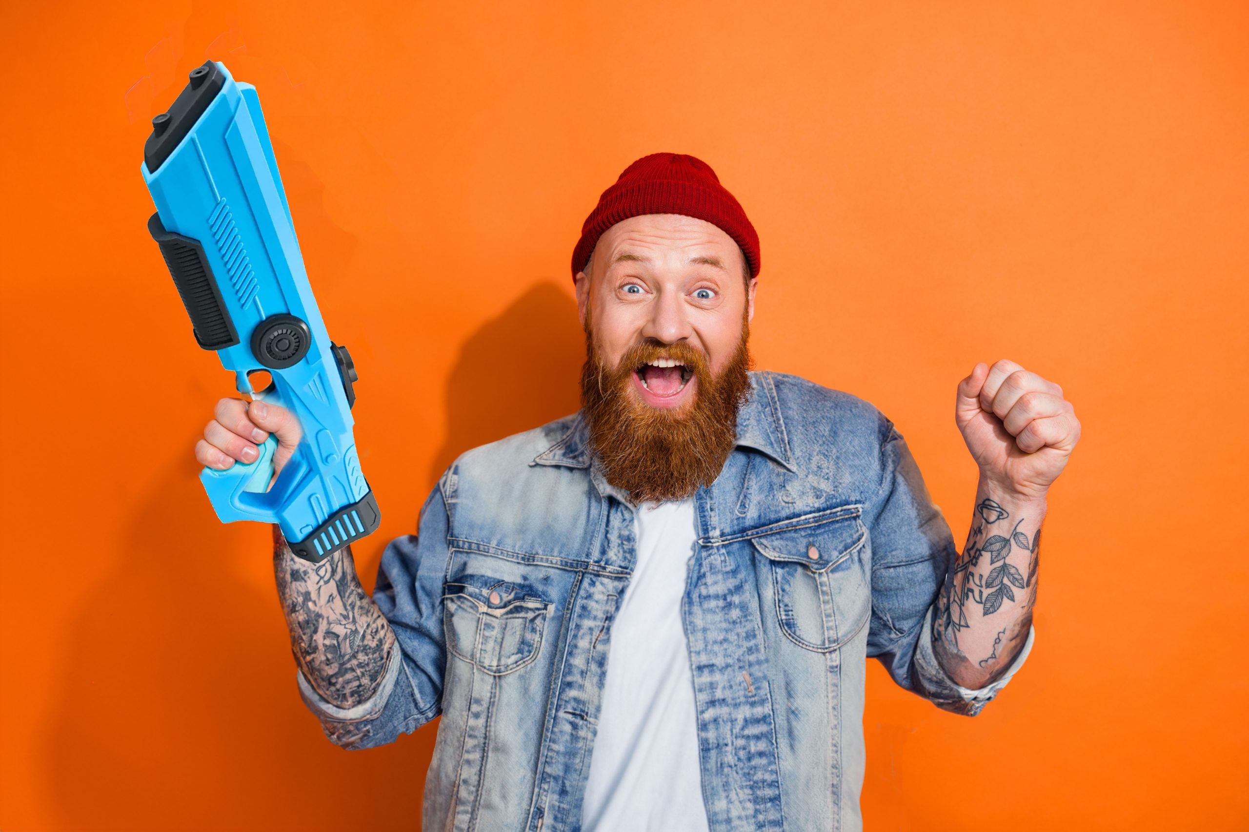 A cheerful man with a red beanie and a beard is holding a toy water gun with a look of mock excitement on an orange background
